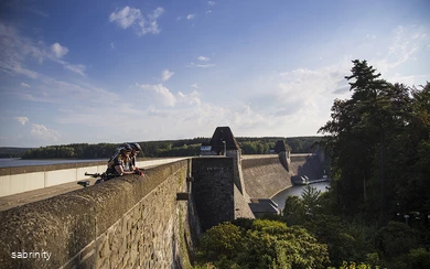 Radfahrer Staumauer Möhnesee (5).jpg Weitblick über die Möhnetalsperre mit Turm, Stausee und umliegender Waldlandschaft