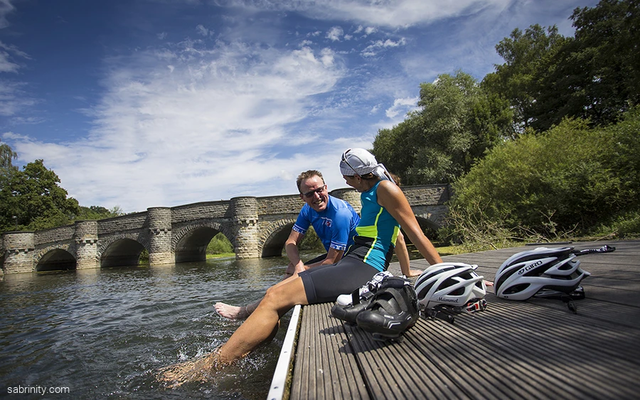Radfahrer plantschen im Wasser Kanzelbrücke Möhnesee (2).jpg Zwei Personen sitzen auf einem Steg am Möhnesee und kühlen die Füße im Wasser, die Kanzelbrücke aus Stein im Hintergrund.