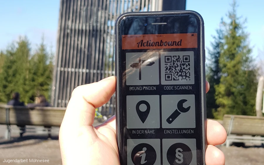 Hand hält ein Smartphone mit der geöffneten App „Actionbound" vor dem Möhnesee-Turm.